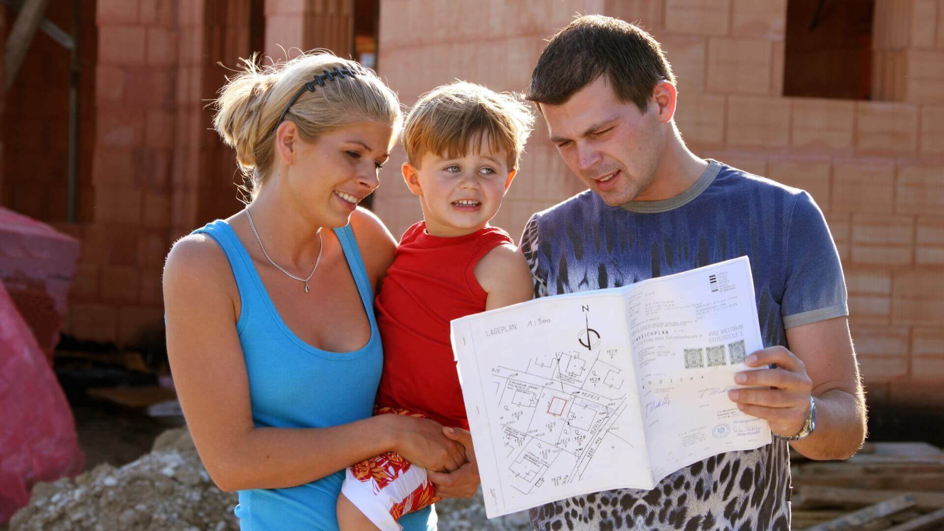 Junge Familie mit Bauplan vor einem Rohbau in Ziegelbauweise – Symbol für Hausbau mit Binder Bau.